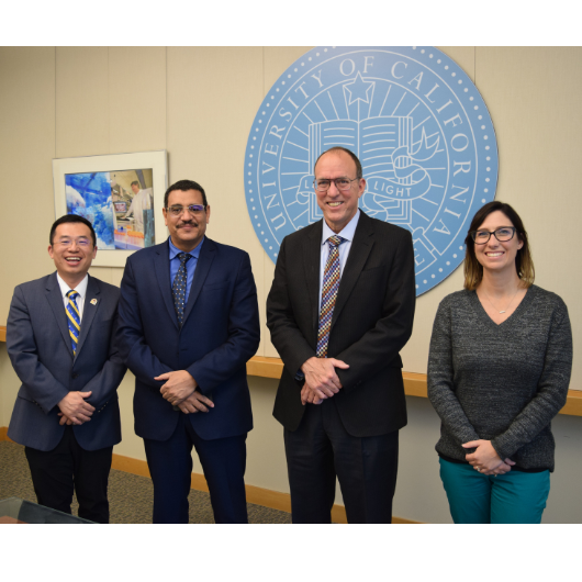 group of people, three men and one women, stand in front of UCR seal on wall